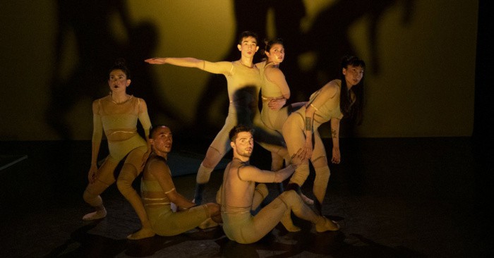 A group of dancers gathered on a dark stage with their shadows projecting on a wall behind them