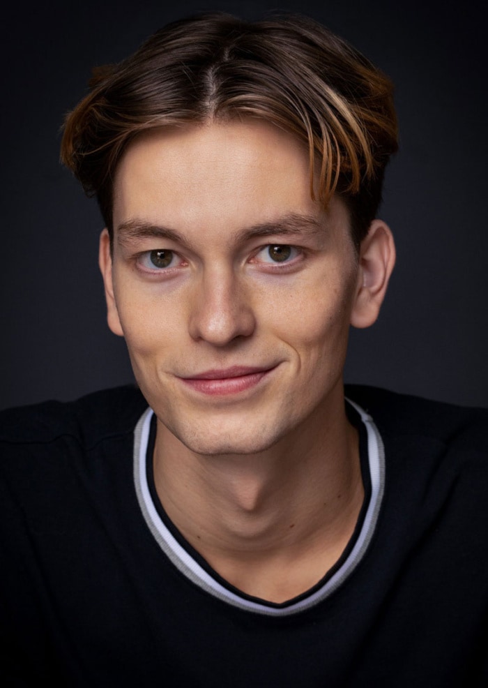 A headshot of Alexander Carroll-Cabanes wearing a dark sweater in front a dark background