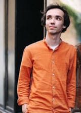 Adam Balogh in an orange button up shirt leaning against a wall