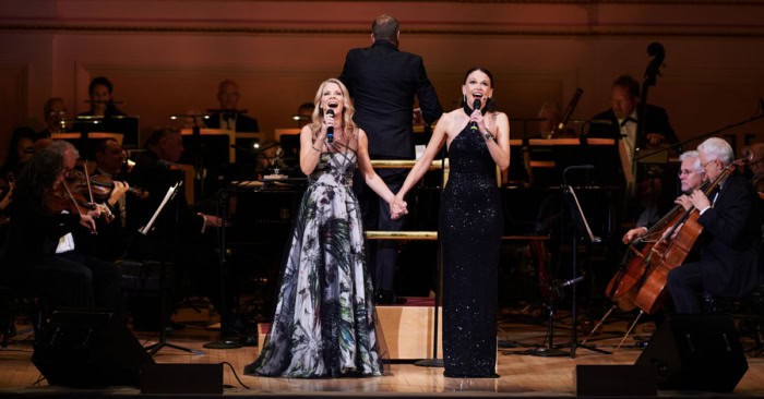 Sutton Foster and Kelli O'Hara singing on stage with an orchestra